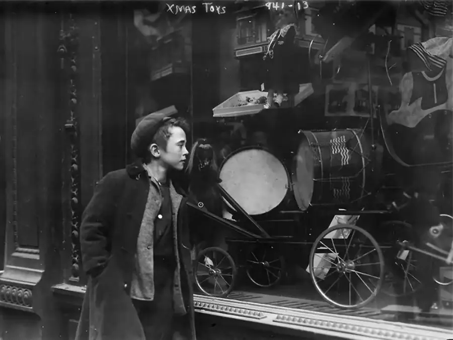 A young boy in a coat and cap gazes longingly into a store window displaying toy drums, a doll, and a toy stroller. The scene appears old-fashioned and festive, with reflections in the glass.