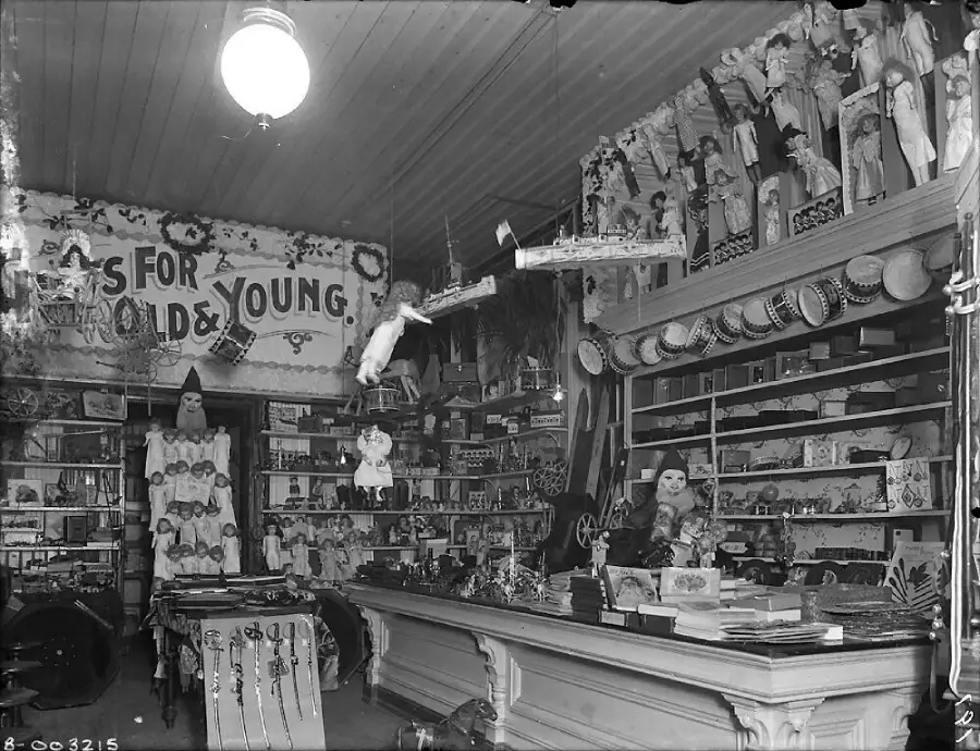 A vintage toy store is filled with dolls, toys, games, and hats. Shelves and counters display an array of items, with decorations hanging from the ceiling and a sign partially reading “FOR OLD & YOUNG.”