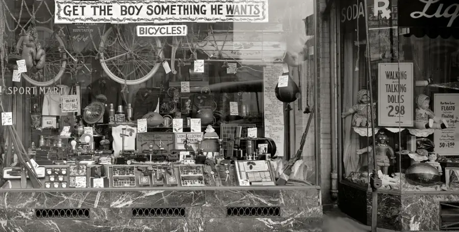A vintage store window displays toys, bicycles, sports equipment, and signs, including one reading “GET THE BOY SOMETHING HE WANTS.” Price tags and handwritten notes are visible among the items.