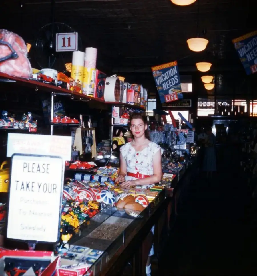 A woman stands behind a counter filled with toys and trinkets in a vintage store. A sign reads, “Please take your purchase to nearest salesgirl.” Hanging signs advertise “Vacation Needs.”