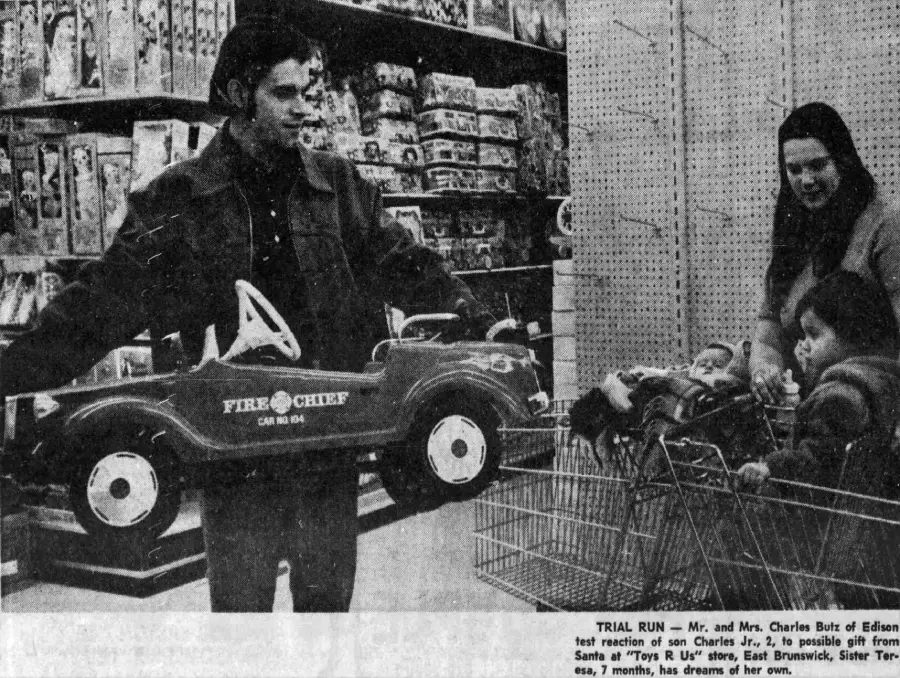 A man holds a red toy fire truck in a toy store while a woman and two children sit in a shopping cart, surrounded by shelves of toys.