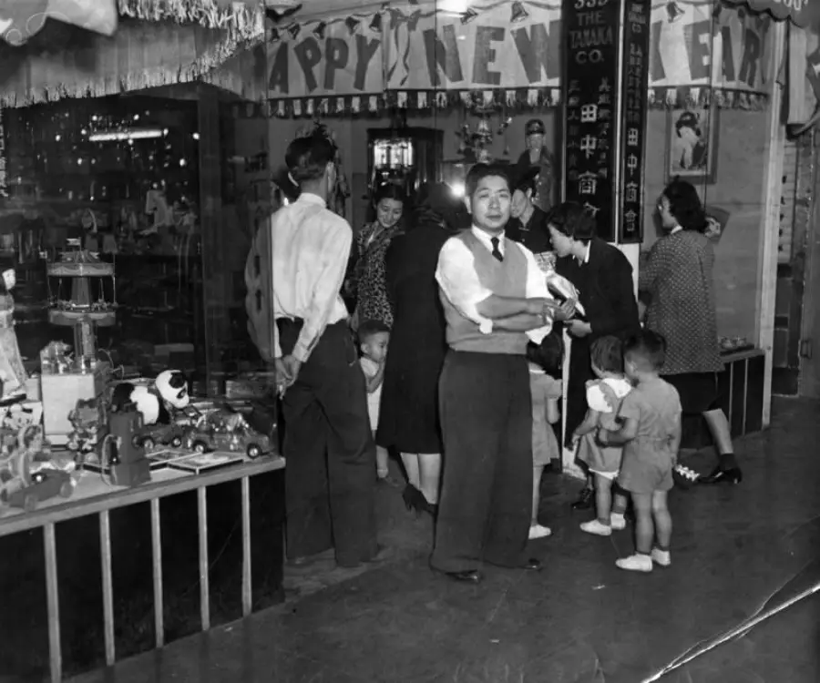 Black-and-white photo of adults and children celebrating New Year’s, with toys displayed on a table and “Happy New Year” banners hanging above in a decorated indoor setting.
