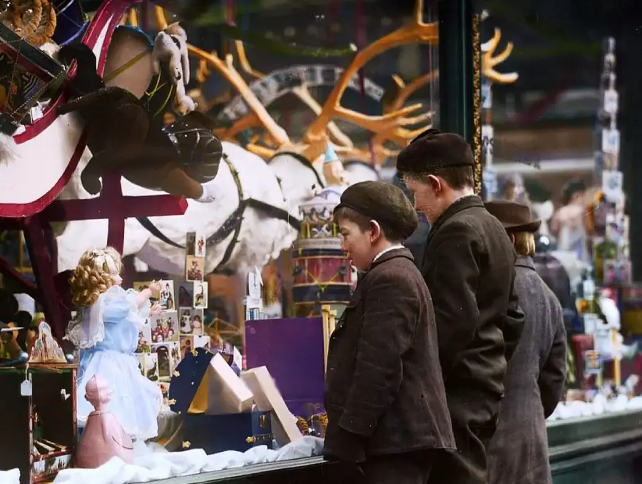 Three boys in old-fashioned clothing gaze through a store window filled with toys, including a doll in a blue dress, a rocking horse, and stacked blocks, with festive decorations displayed inside.