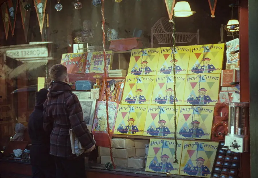 Two people look into a toy store window displaying many “Hop Ching” board games, colorful decorations, toys, and hanging flags. Warm lights illuminate the lively holiday scene.