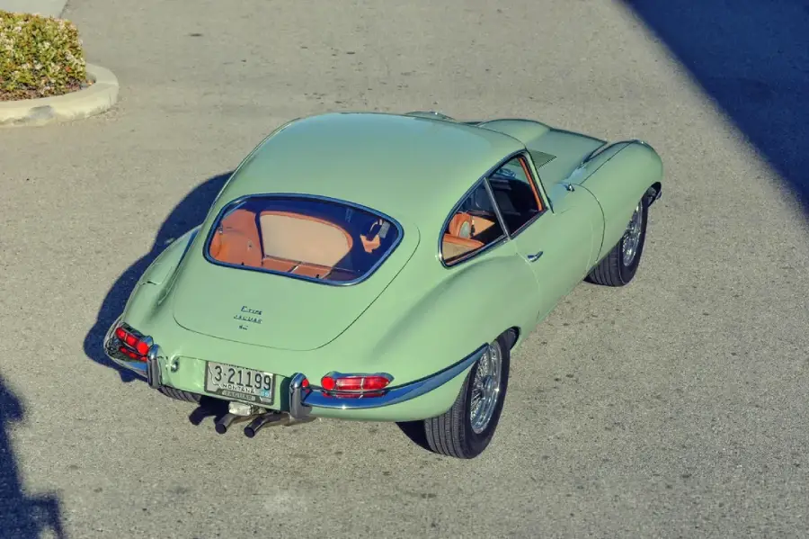 A light green classic coupe car with tan interior is parked on a sunlit street, seen from above at a rear angle. The car has chrome bumpers and vintage styling.