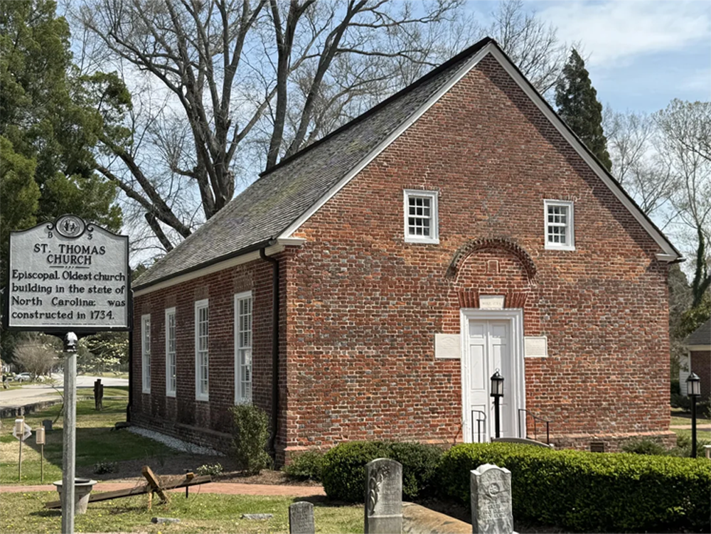A small, historic red brick church with white trim and arched windows, surrounded by gravestones. A sign in front reads “St. Thomas Church, Episcopal, Oldest church building in North Carolina, constructed in 1734.”