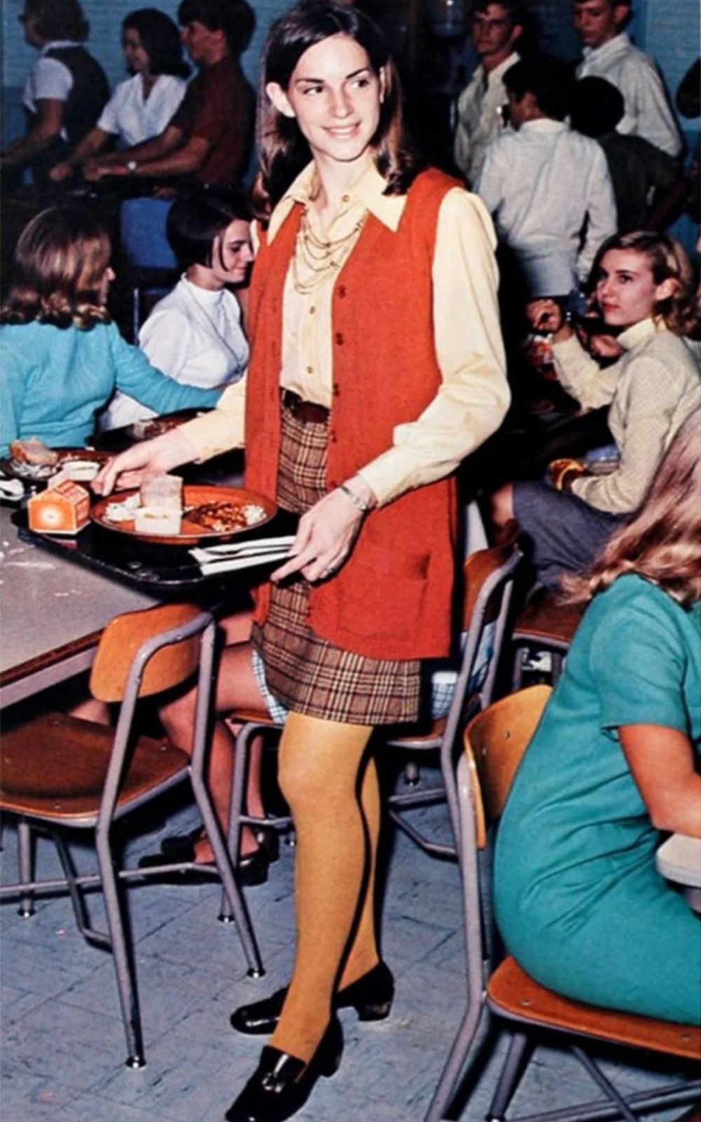 A young woman in 1970s fashion carries a lunch tray in a busy cafeteria, wearing a plaid skirt, orange vest, yellow tights, and a beige blouse. Other students sit and chat at tables in the background.