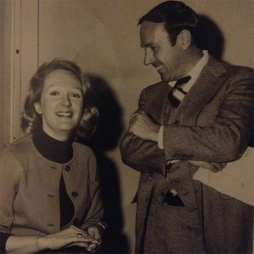 A woman smiles at the camera while a man in a suit stands beside her, looking at her and smiling. The photo is in black and white and appears vintage.
