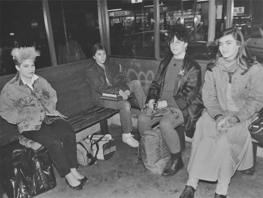 Four young people sit on a bench at a bus stop at night, wearing 1980s-style clothing and jackets, with bags on the floor beside them. Storefronts and streetlights are visible through the windows behind them.