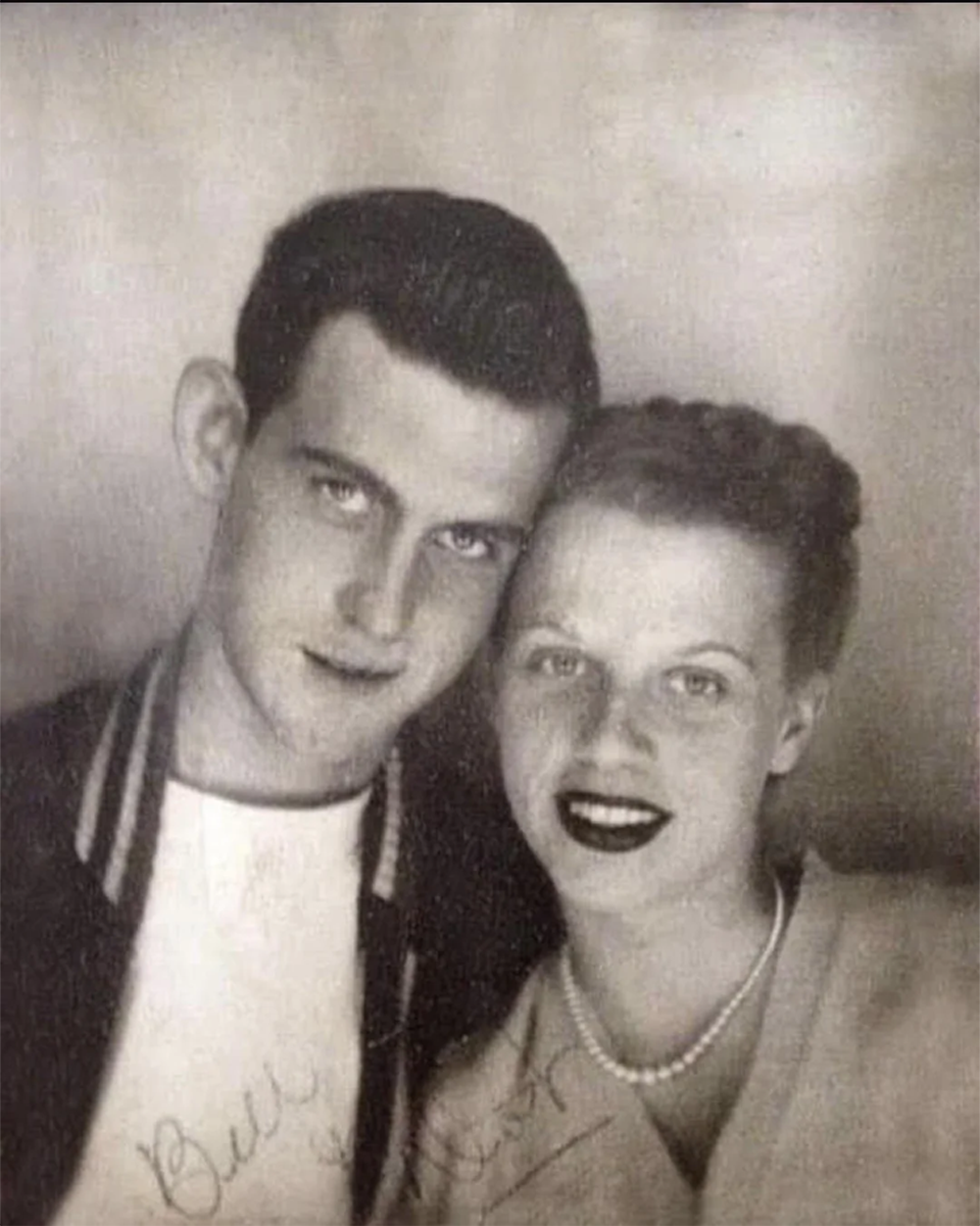 A black-and-white photo of a young man in a jacket and a young woman in a sweater with a pearl necklace, smiling and leaning close together. Both have autographs written on the lower part of the image.