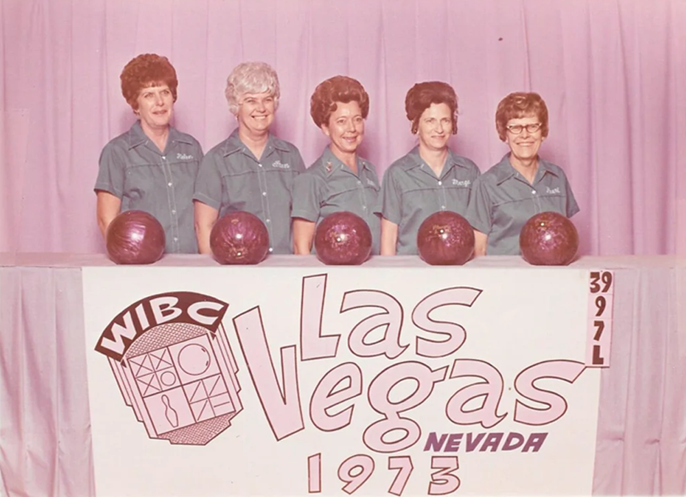 Five women in matching teal shirts stand behind a table with bowling balls. The table displays a large sign reading “WIBC Las Vegas Nevada 1973.” The background is pink with a curtain.
