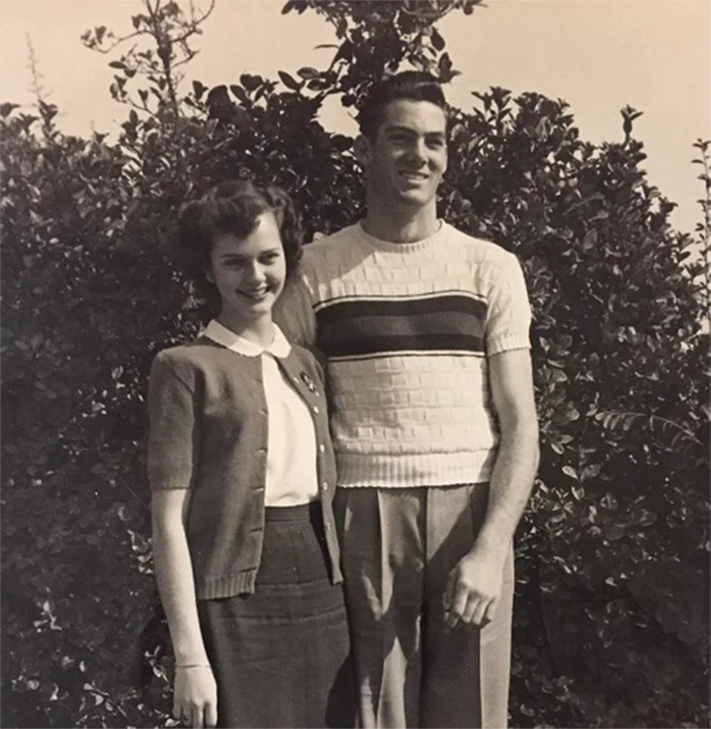 A young woman and man stand close together, smiling, in front of leafy bushes. The image appears vintage, in black and white.