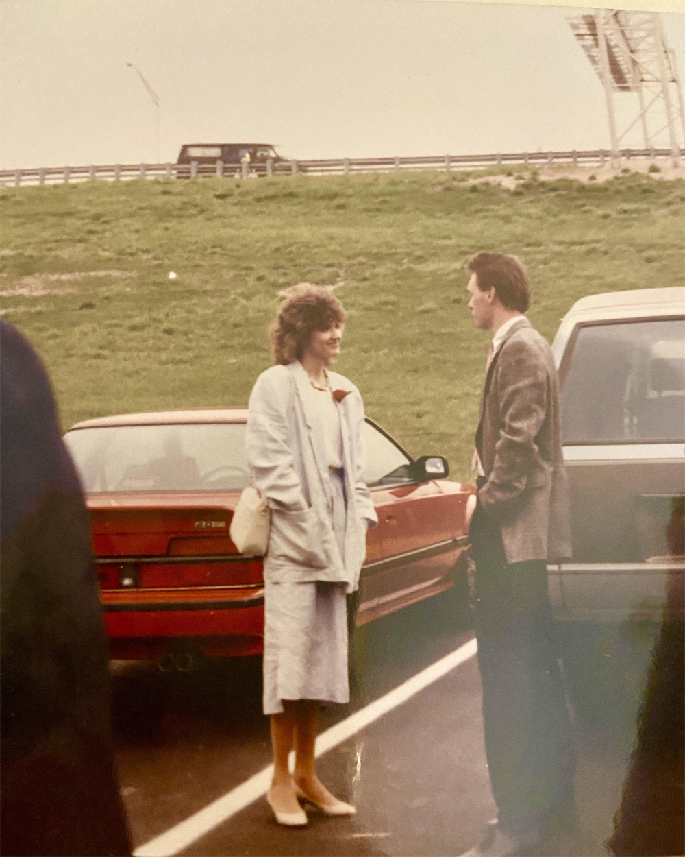 A woman and a man stand talking in a parking lot near a red car. The woman is wearing a light blue outfit and the man is in a suit. A road and grassy hill are visible in the background.
