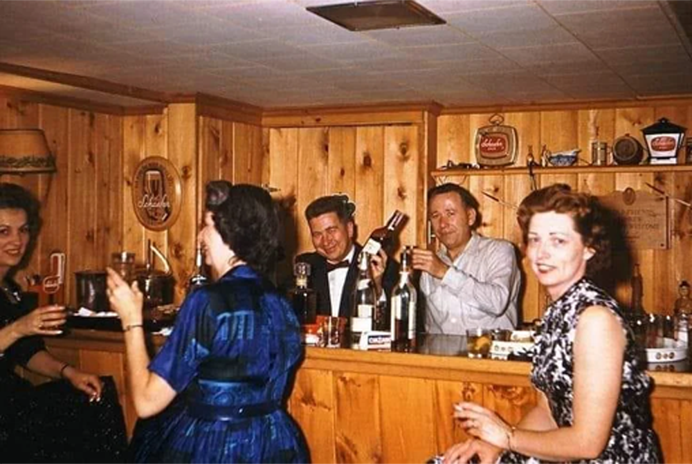 Four people sit at a wooden home bar, holding drinks and smiling. Two men stand behind the bar with bottles, and two women in vintage dresses sit in front, facing the camera. The setting has a cozy, retro atmosphere.