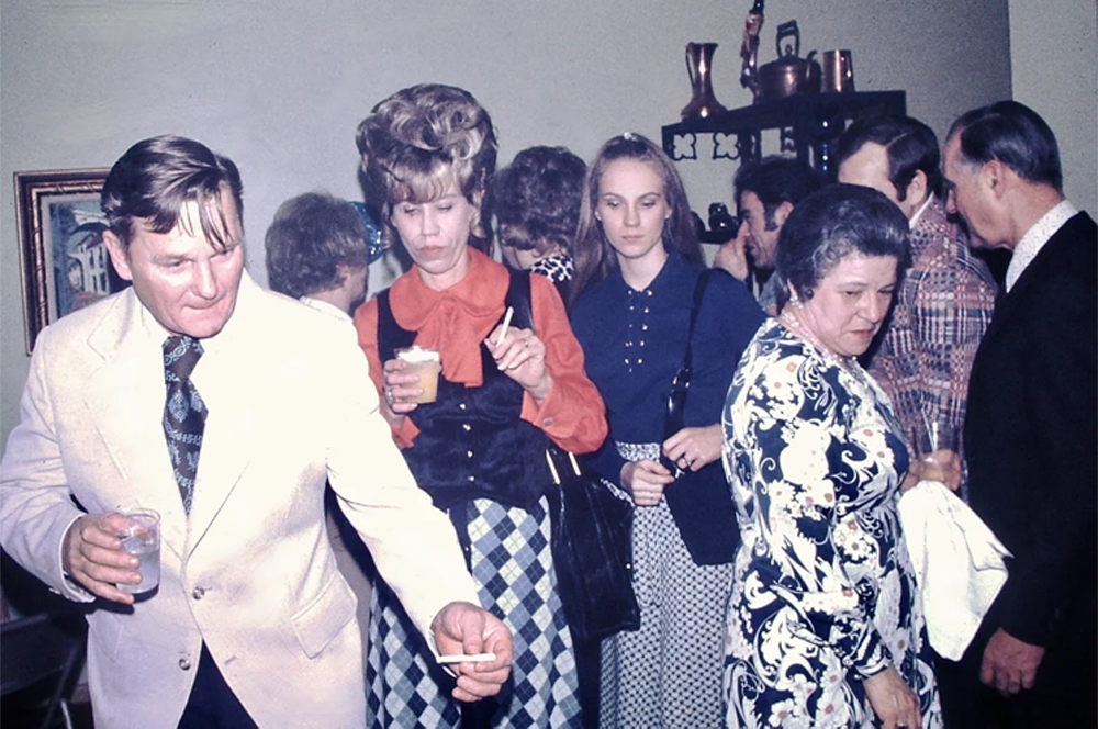 A crowded indoor party scene from the 1970s showing men and women in vintage clothes, some holding drinks, one man reaching out with a cigarette, and others talking in a lively atmosphere.