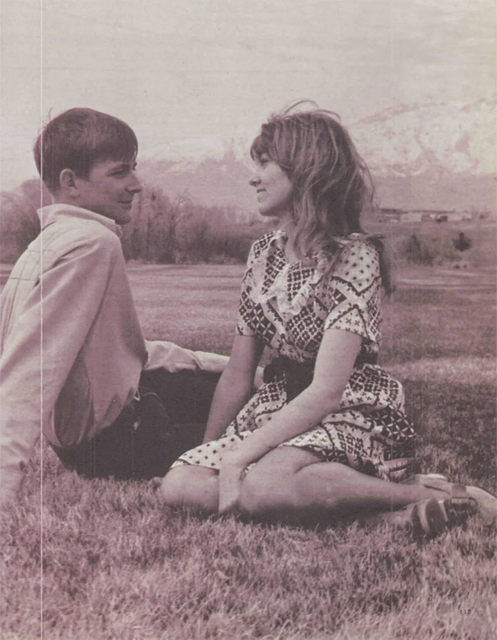 A young man and woman sit on grass facing each other, smiling, with mountains and trees in the background. The image is in black and white, creating a vintage feel.