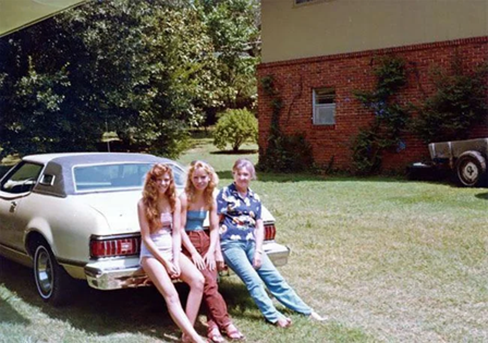 Three young people sit on the bumper of a vintage beige car parked on a sunny lawn. A brick house and trees are in the background, with another vehicle partially visible on the right.
