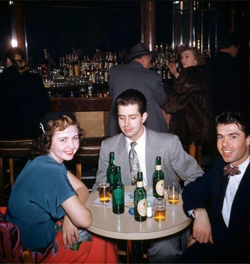 Three people sit at a small round table with bottles and glasses, smiling at a bar. Two women and a man are seen at the bar in the background. The setting appears vintage.