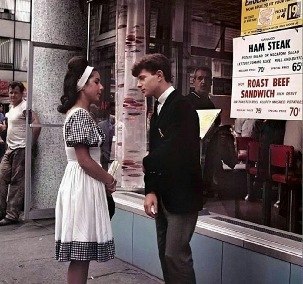 A young woman in a checkered dress and a young man in a suit stand talking outside a diner with a menu in the window. People walk by on the busy city street.