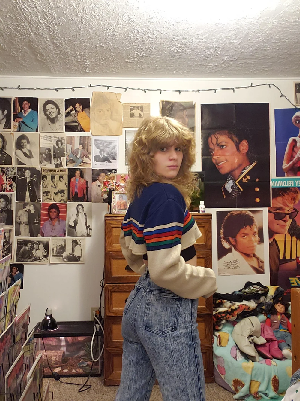 A person with wavy blonde hair, wearing a striped sweater and acid-wash jeans, stands in a bedroom filled with Michael Jackson posters and memorabilia, looking over their shoulder at the camera.