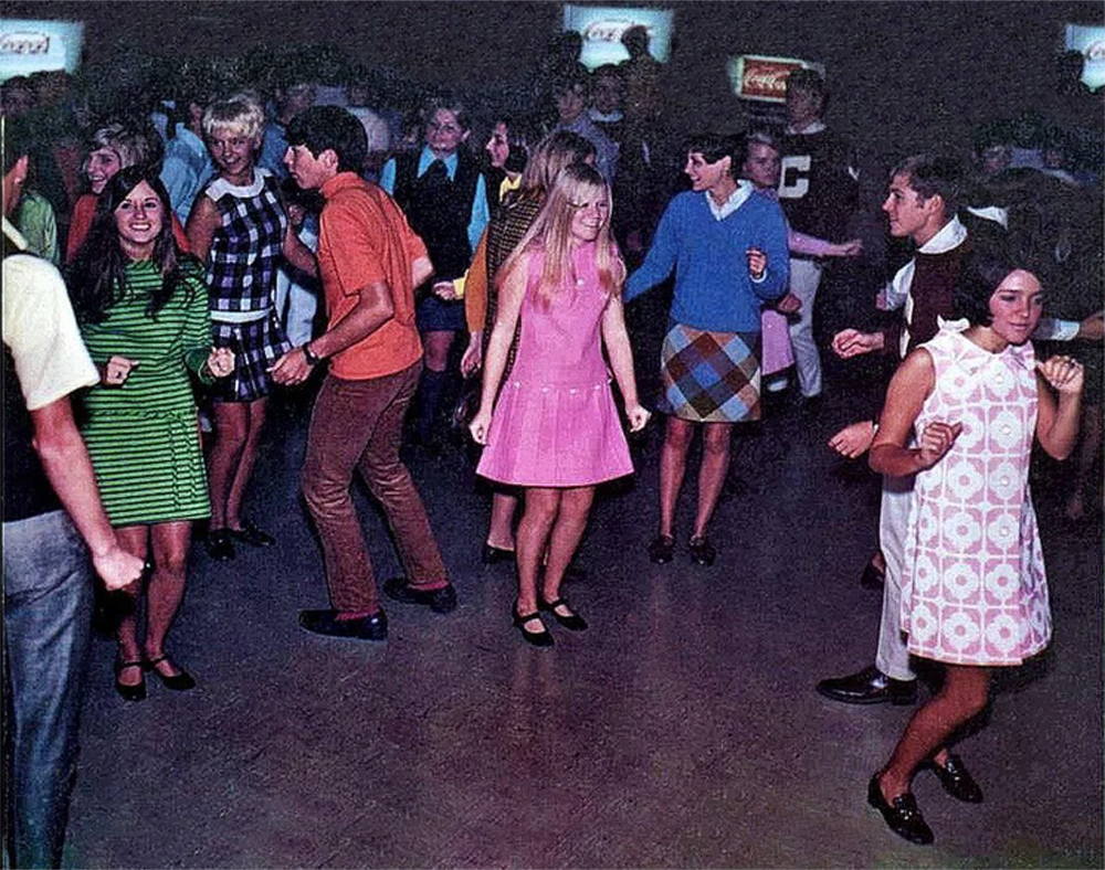 Group of young people in colorful 1960s outfits dance at a crowded indoor event. The floor is open, and everyone appears to be enjoying themselves, moving to the music.