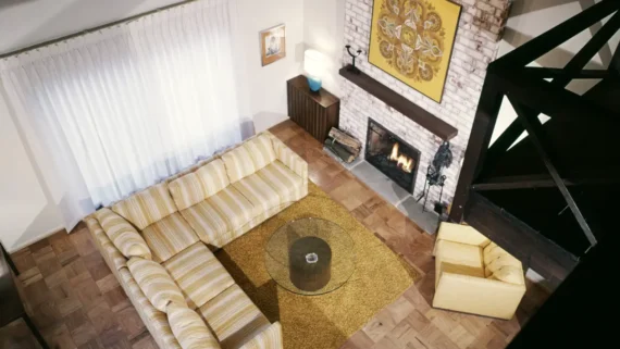 Aerial view of a cozy living room with a striped sectional sofa, yellow armchair, glass coffee table, rug, fireplace, and wall art above the mantel.