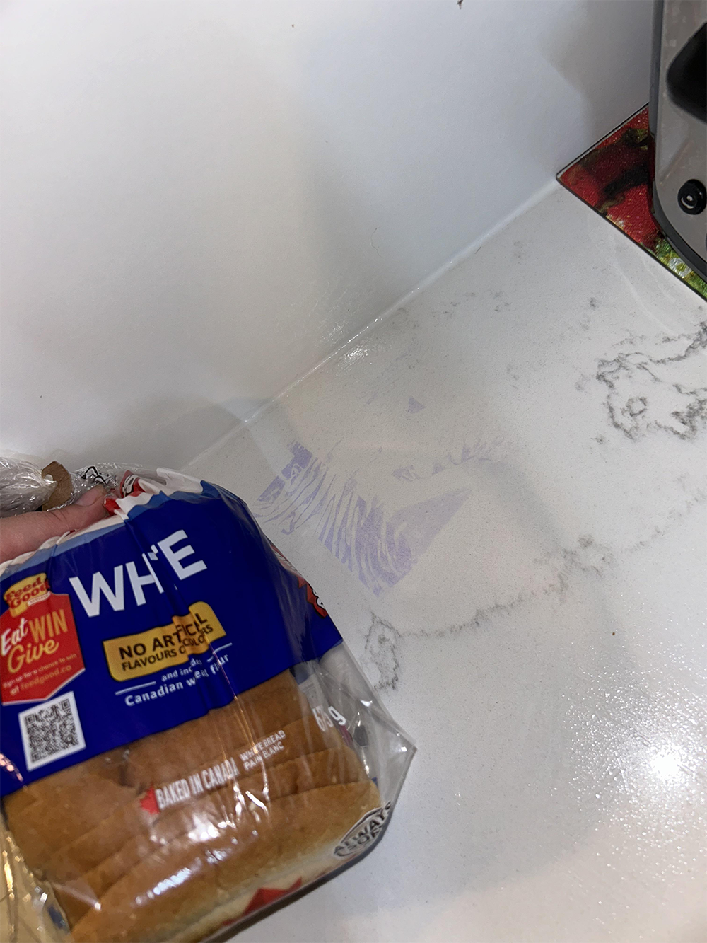 A loaf of white bread in blue packaging rests on a white marble countertop under bright lighting. Part of a toaster and a red object are visible in the top right corner.