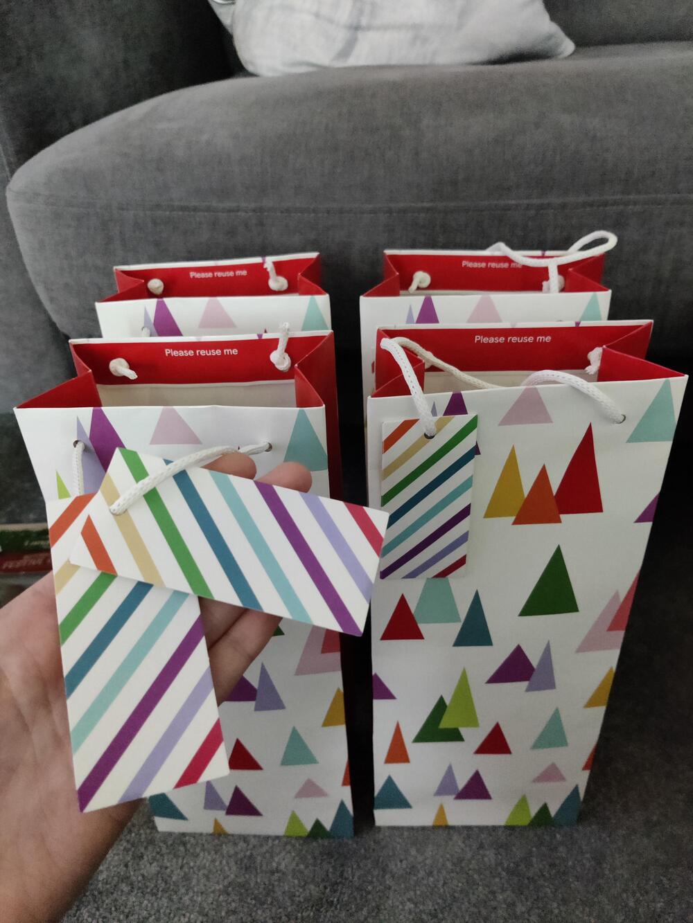 Four white gift bags with colorful triangle patterns and striped tags are held in front of a gray couch. A hand is holding one bag closer to the camera, displaying the attached striped tag.