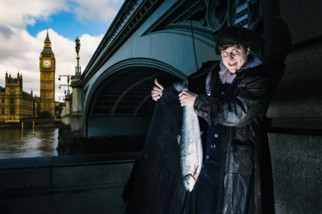 A person in historical costume stands by a river in London, near Big Ben and a bridge, smiling while holding a large fish under their coat.