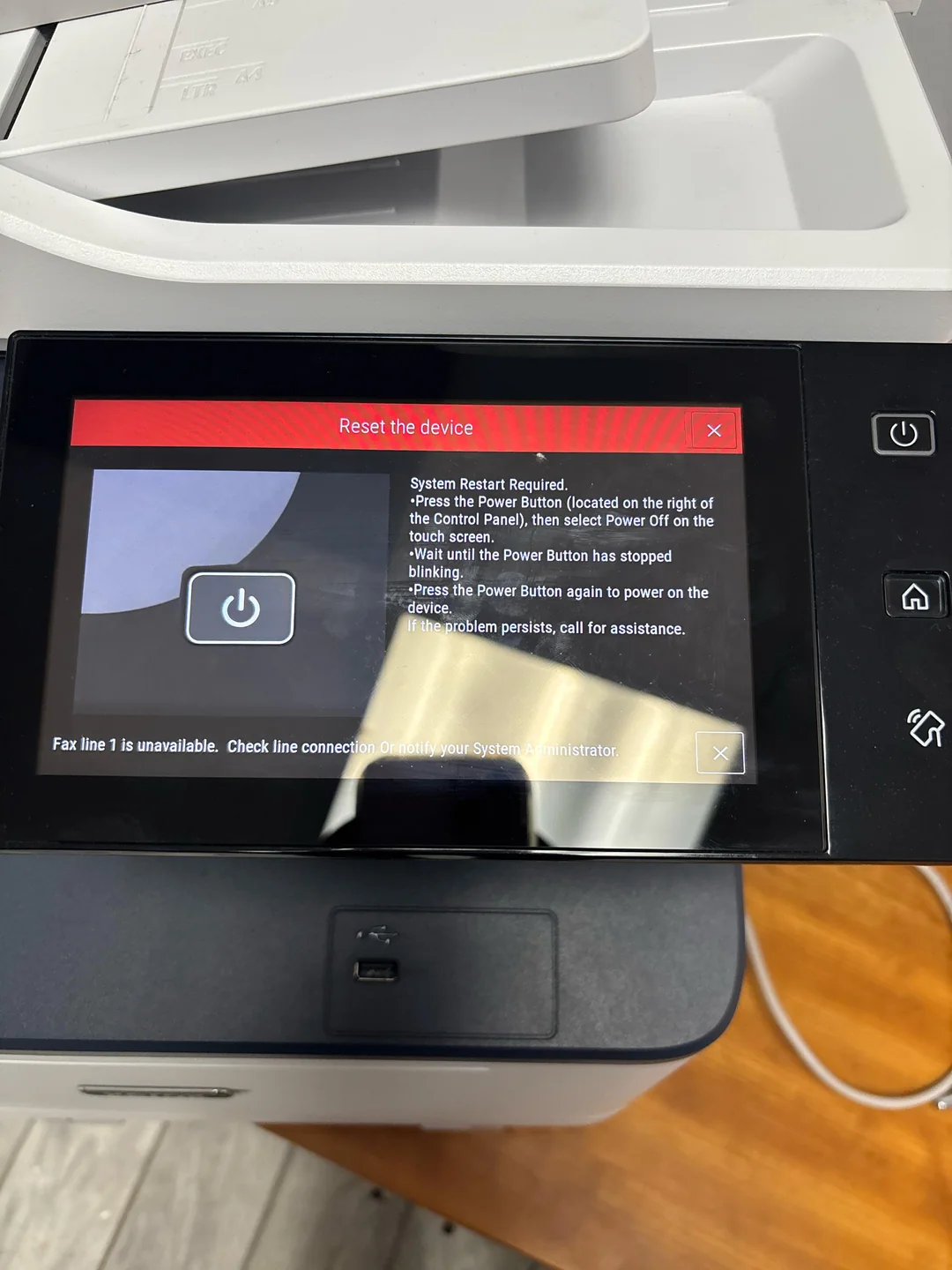 Printer touchscreen displays a “Reset the device” message with power cycle instructions; nearby, a corner of the printer and its paper tray are visible. A warning about fax line connection is shown at the bottom.
