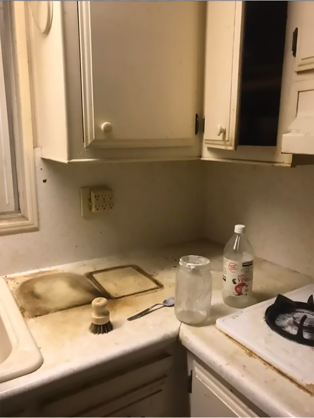A cluttered, dirty kitchen corner with stained countertops, a scrubbing brush, a spoon, an empty jar, and a bottle of vinegar beside a gas stove. Cabinets above are slightly open.