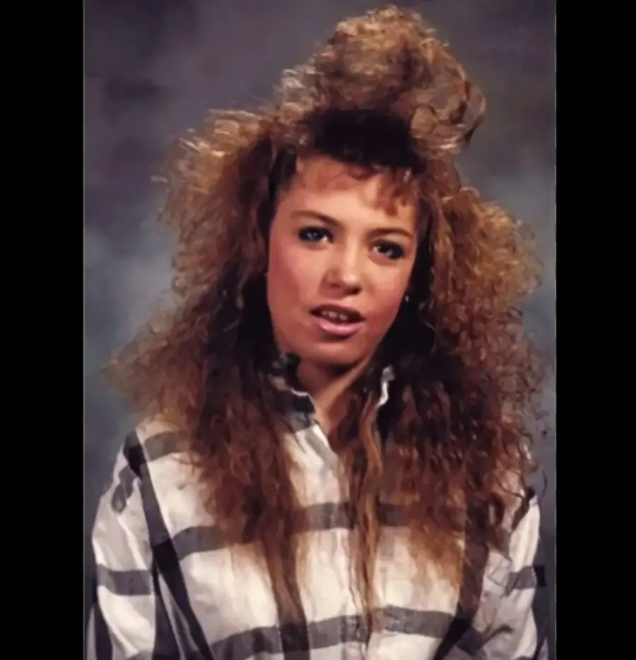 Young woman with voluminous, teased curly hair and a plaid shirt poses for a studio portrait against a gray background, giving a slight smile.
