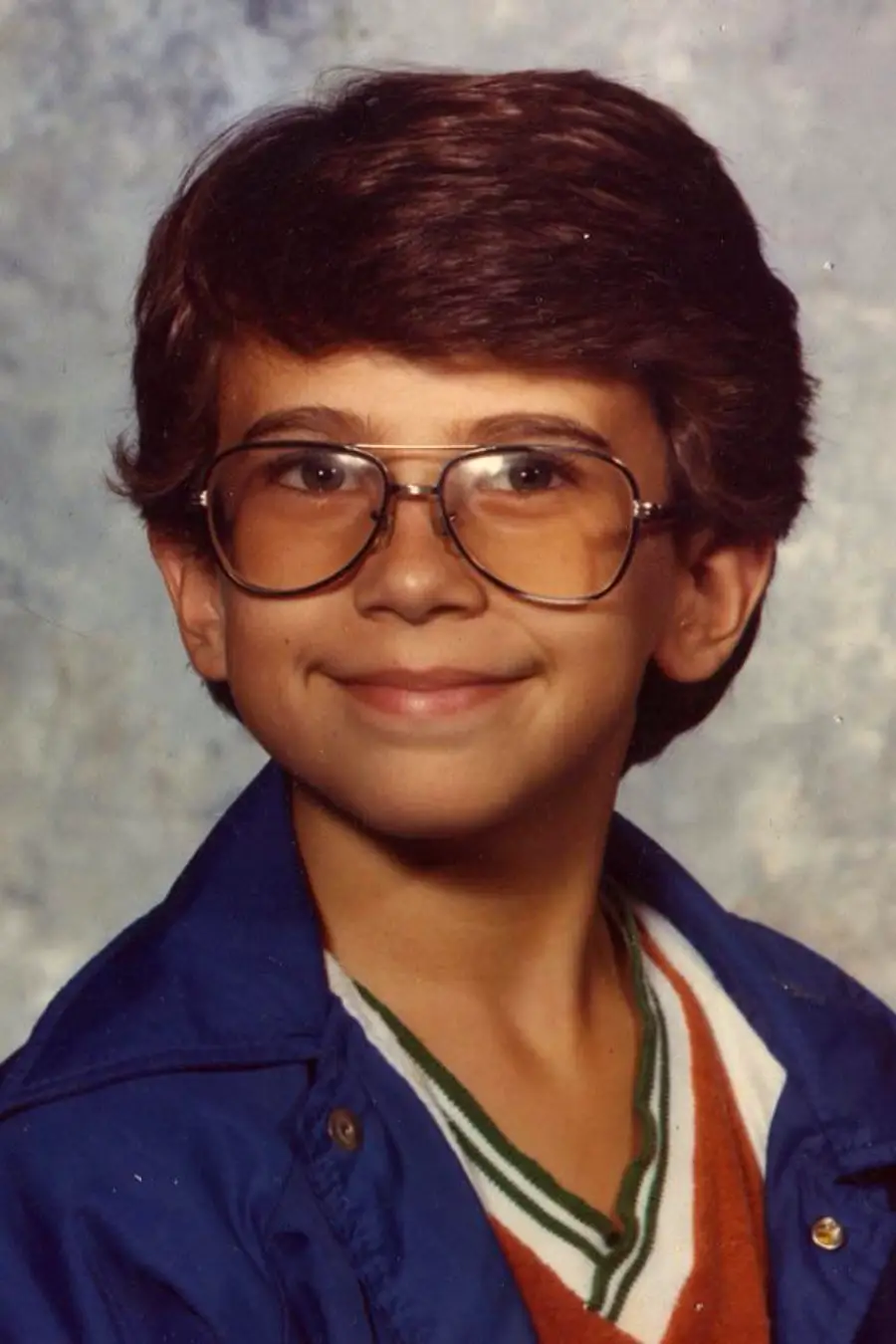A young boy with brown hair and large glasses smiles at the camera, wearing a blue jacket over an orange shirt with a white and green collar, in front of a mottled studio backdrop.