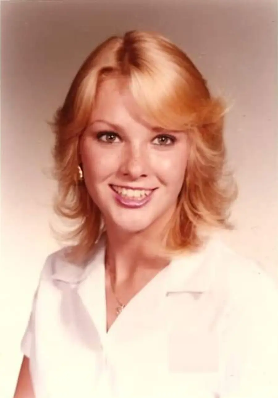 A young woman with blonde hair styled in loose waves smiles at the camera, wearing a white collared shirt and small earrings, against a plain, light-colored background.