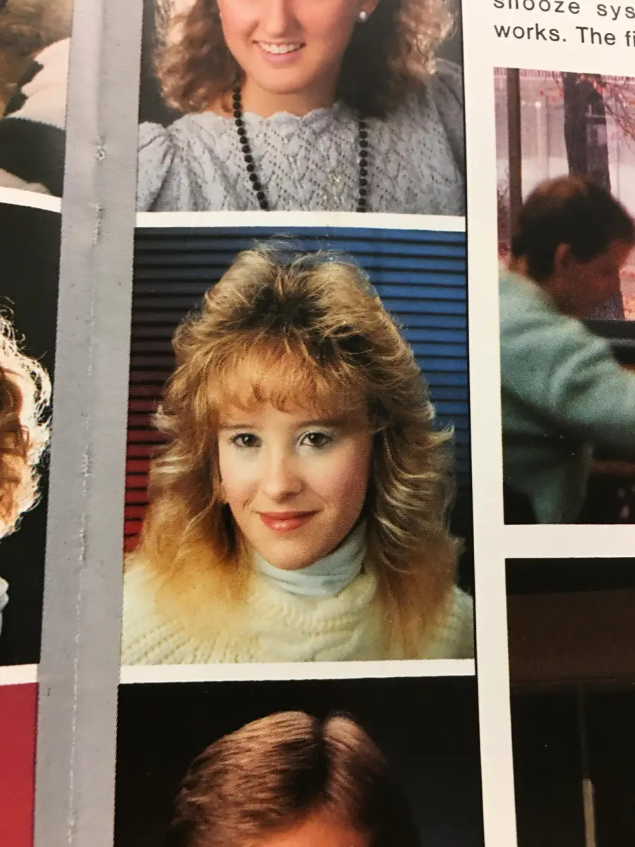 A young woman with blonde, feathered hair wearing a white turtleneck sweater poses for a school portrait against a red and blue striped background.