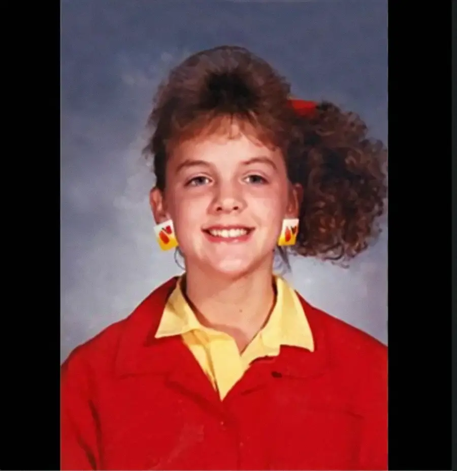 Smiling girl with curly side ponytail, colorful bow, red jacket, and yellow shirt, wearing large geometric earrings, posing for a school photo against a gray background.