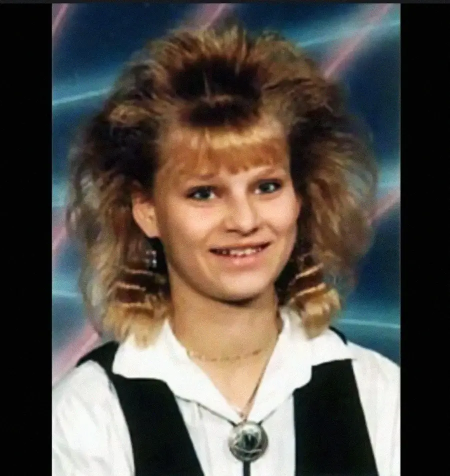 Smiling person with teased, voluminous blonde hair, wearing a white shirt, black vest, and bolo tie. The background features a blue backdrop with pink diagonal streaks, resembling a retro school photo style.