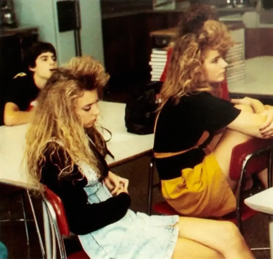Three students sit in a classroom with desks and a stack of books in the background. Two girls in front have big 1980s-style hair and are wearing casual outfits. They appear to be listening or waiting.