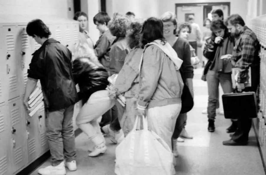 Black and white photo of students crowded in a school hallway, some opening lockers and others talking or walking. Most wear casual 1980s clothing, including jackets and sneakers.