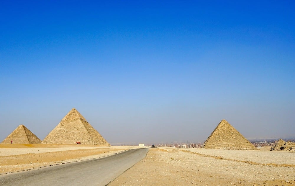A paved road leads toward three large pyramids under a clear blue sky, with desert sand and a distant city visible on the horizon.