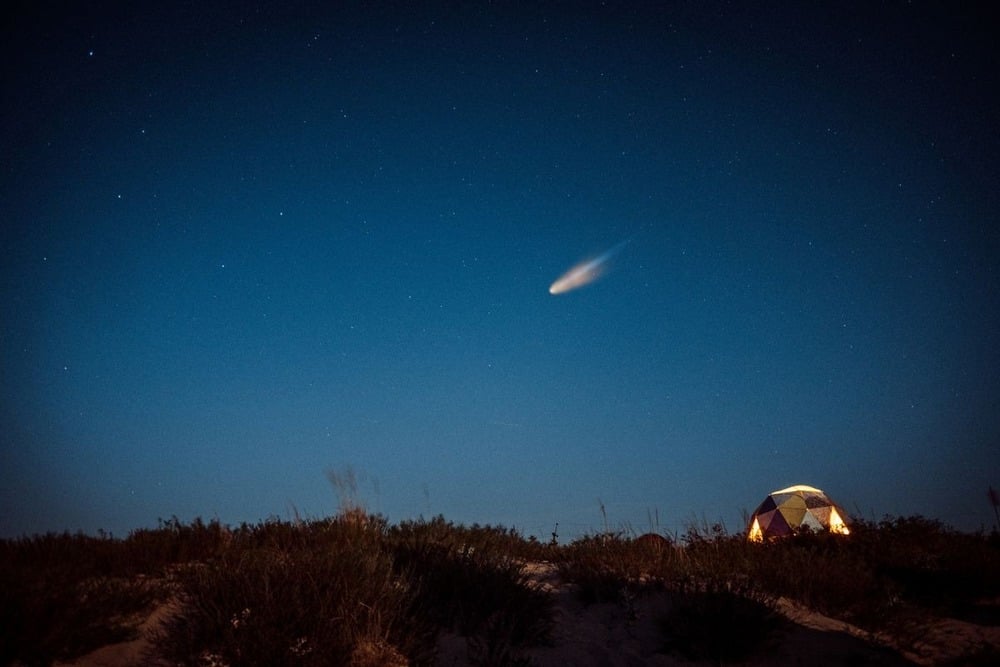 A glowing tent sits on a grassy hill under a starry night sky, with a bright comet or meteor streaking across above.