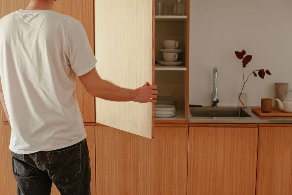 A person opens a wooden kitchen cabinet, revealing neatly stacked plates and cups. Nearby is a sink, countertop items, and a vase with dark leaves. The scene is warm and minimalistic.