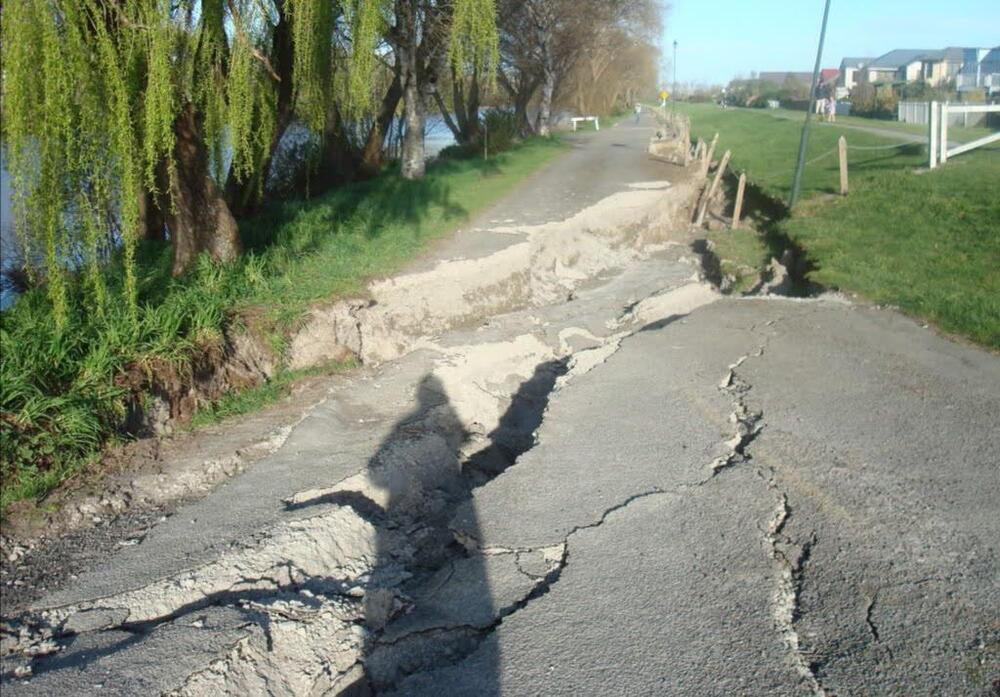 A cracked and damaged riverside path with deep fissures in the asphalt, bordered by green grass, trees, and nearby houses in the background.
