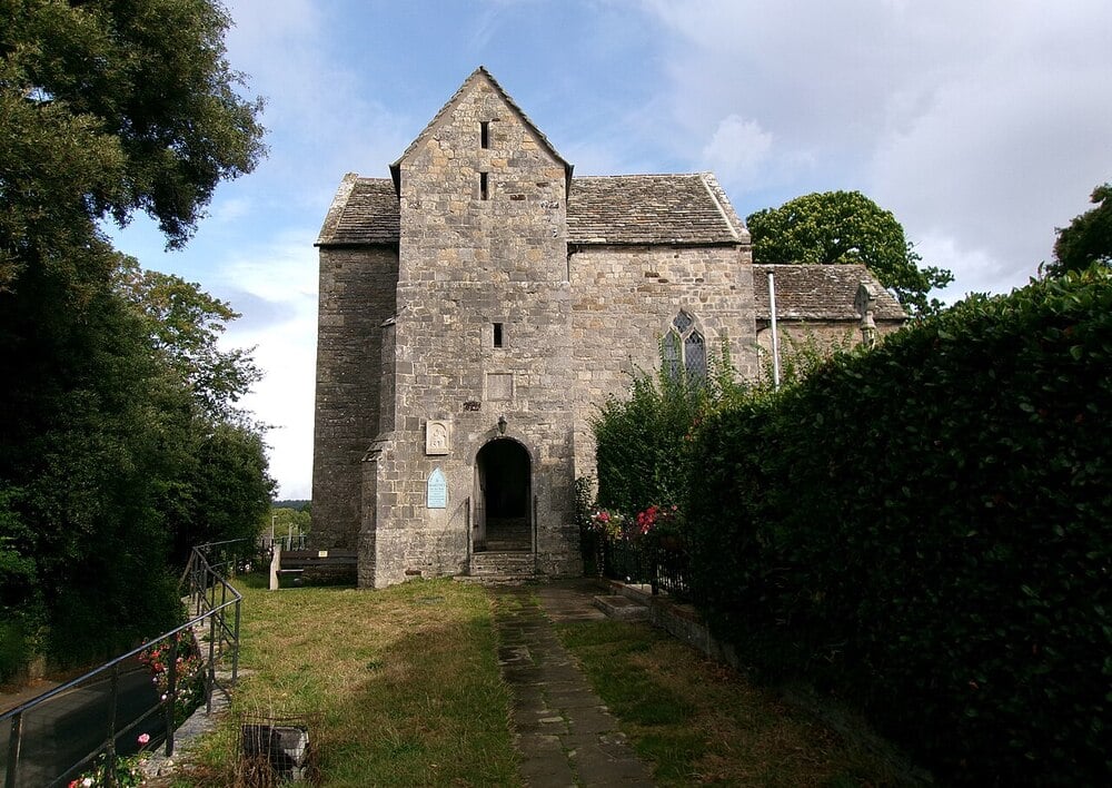 A small, old stone church with a steep roof, arched entryway, and narrow windows, surrounded by greenery and a grassy pathway leading to the entrance under a partly cloudy sky.
