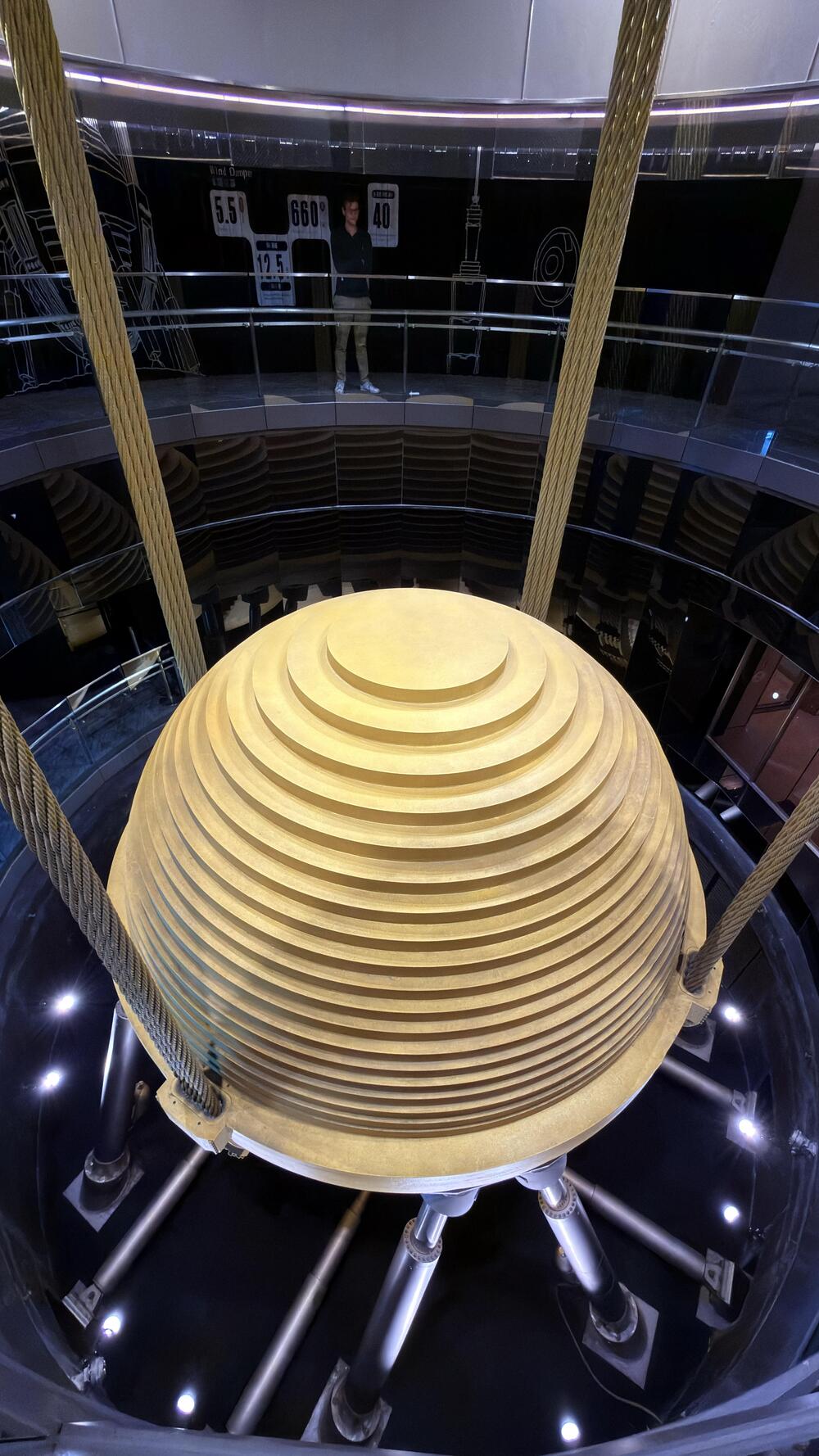 A large, gold, tiered sphere suspended by thick cables inside a glass enclosure, viewed from above, with a person standing on an upper walkway in the background.