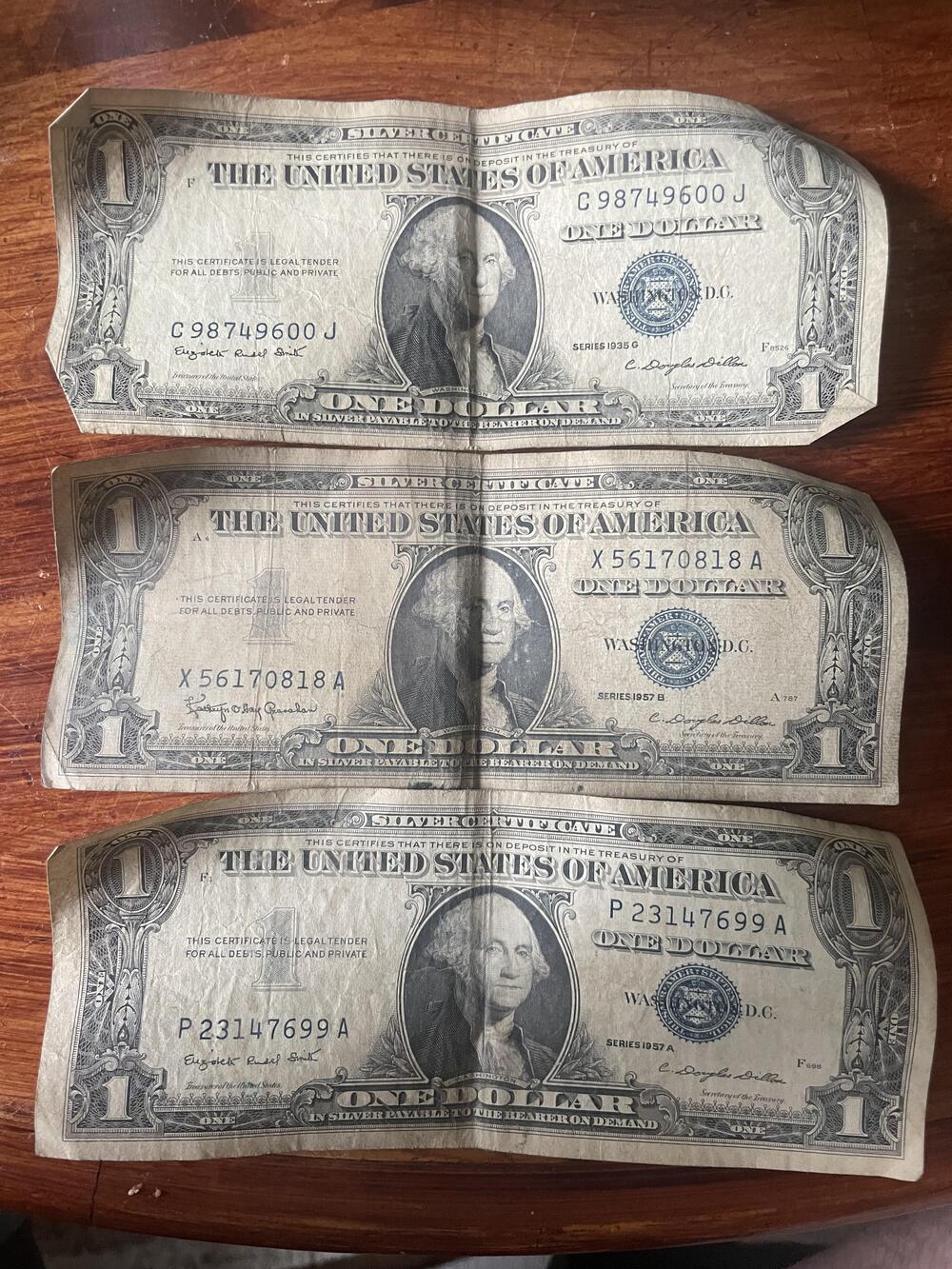 Three worn U.S. one-dollar bills are laid out horizontally on a wooden surface, showing front images of George Washington and visible serial numbers.