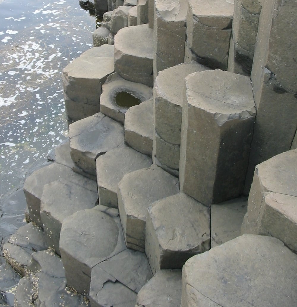 Hexagonal stone columns rise from the shoreline beside the water, forming a natural geometric pattern. Some stones are wet from the waves.