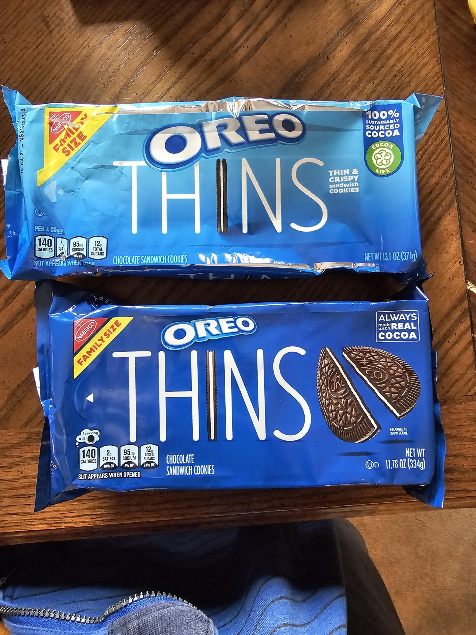 Two packages of Oreo Thins cookies are on a wooden table. The top package is blue and labeled Family Size, and the bottom package is darker blue, also Family Size.
