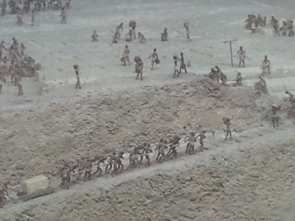 Diorama showing workers moving a large stone block with ropes while others work on a barren, rocky landscape, likely depicting an ancient construction scene.