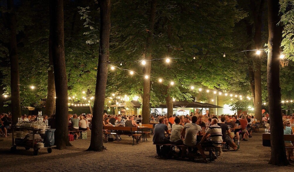 People gather at picnic tables in an outdoor beer garden at night.